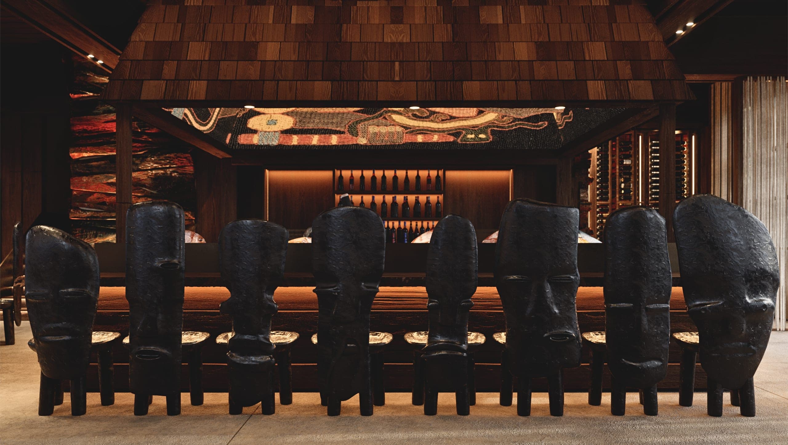 Bamboomat: row of sculptural black stools with carved faces at the bar, under a shingled canopy